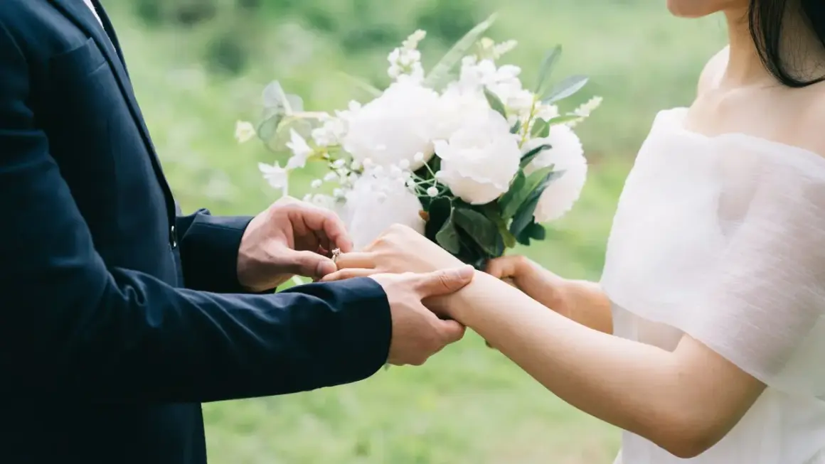 Un homme en costume met une bague à la main d'une femme, bouquet de fleurs blanches. Un poème mariage Baudelaire pour un amour éternel.
