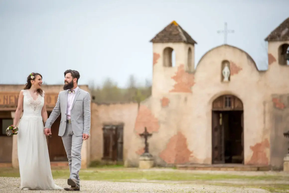Un couple rayonnant, tout juste marié, se promène main dans la main devant une chapelle rustique, évoquant un mariage western.