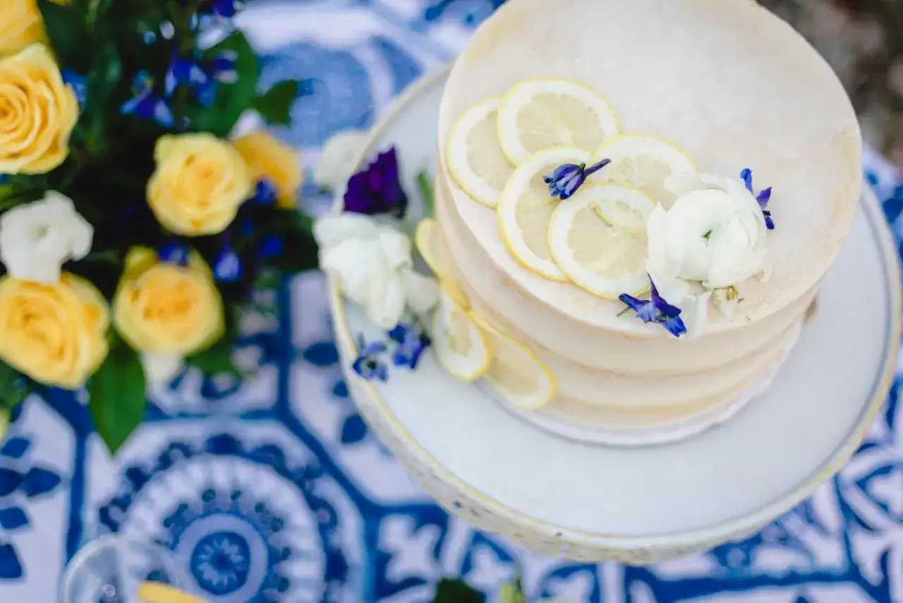 Gâteau de mariage jaune et bleu, décoré de rondelles de citron et de fleurs. Un bouquet de roses jaunes et de fleurs bleues complète la scène.