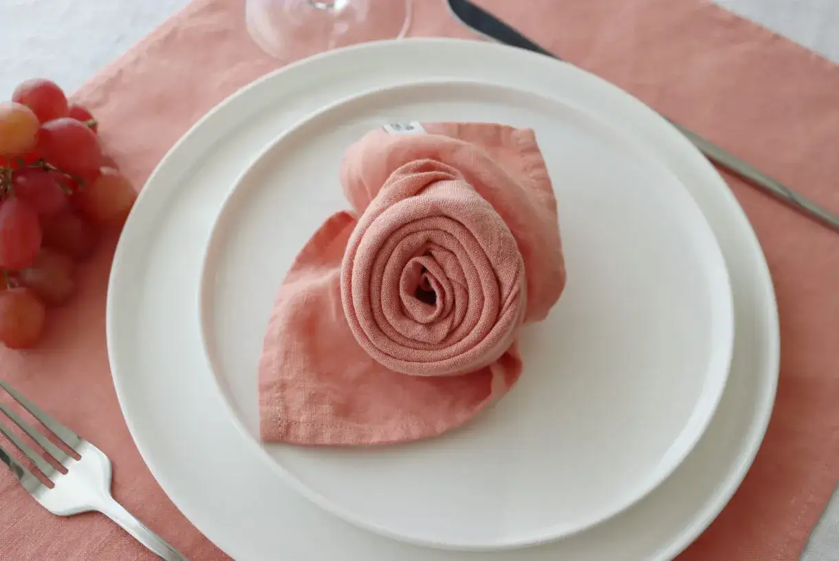 Une serviette rose pliée en forme de rose sur une assiette blanche, un bouquet de raisins rouges à côté. Un pliage serviette original pour une table élégante.