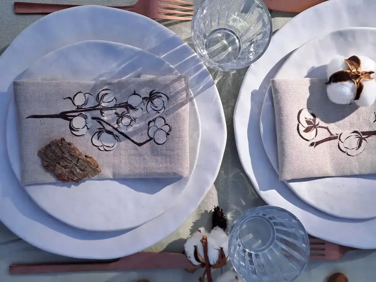 Table dressée pour 1 an de mariage, noce de coton. Serviettes ornées de branches de coton, assiettes blanches, verres texturés et couverts cuivrés.