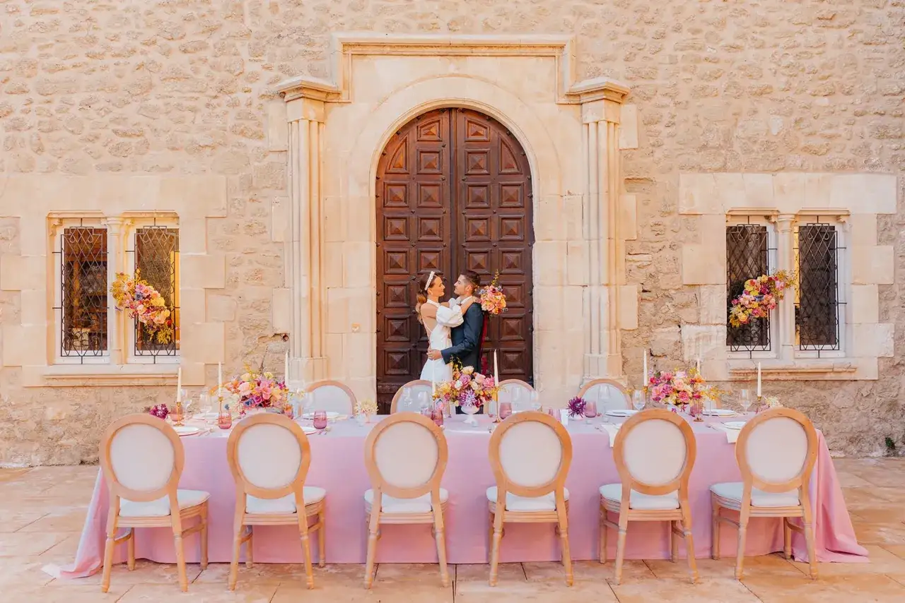 Un couple s'embrasse devant une porte massive, une table dressée pour un festin gourmand, ornée de fleurs roses et de bougies.
