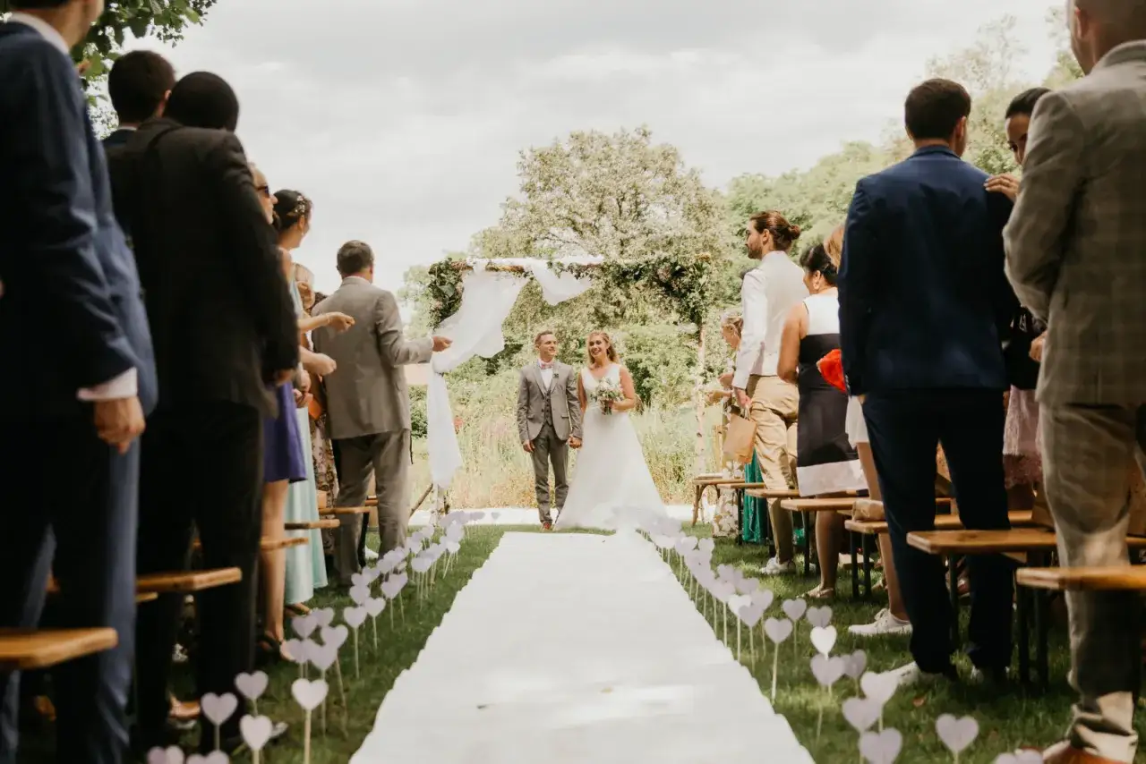 Un couple rayonne sur l'all&eacute;e de mariage ext&eacute;rieur, entour&eacute; d'invit&eacute;s attentifs. Le chemin blanc est bord&eacute; de c&oelig;urs, cr&eacute;ant une atmosph&egrave;re romantique.