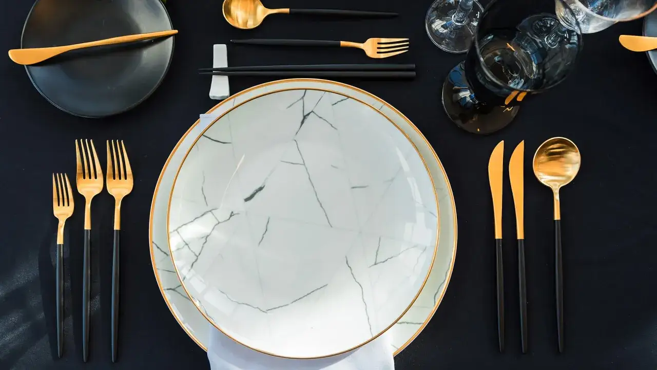 Service à l'anglaise : vaisselle élégante avec des assiettes marbrées, couverts dorés et noirs, et verres à vin sur une nappe noire.