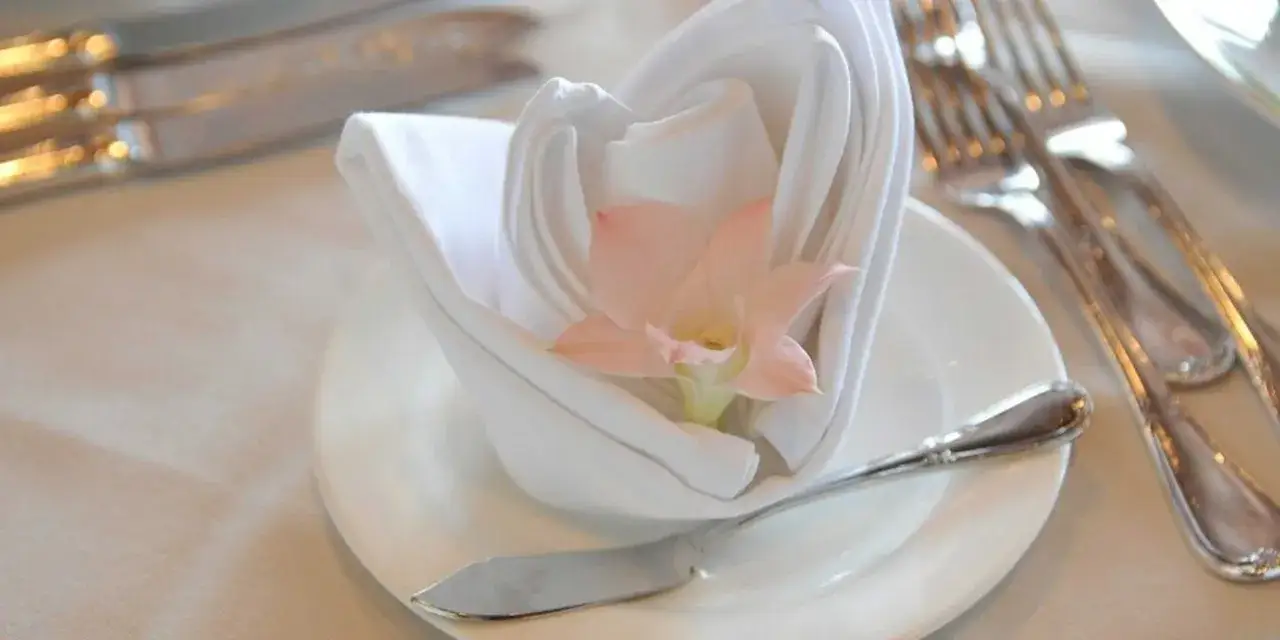 Un &eacute;l&eacute;gant noeud serviette mariage blanc orn&eacute; d'une fleur rose p&acirc;le, sur une assiette avec des couverts argent&eacute;s.