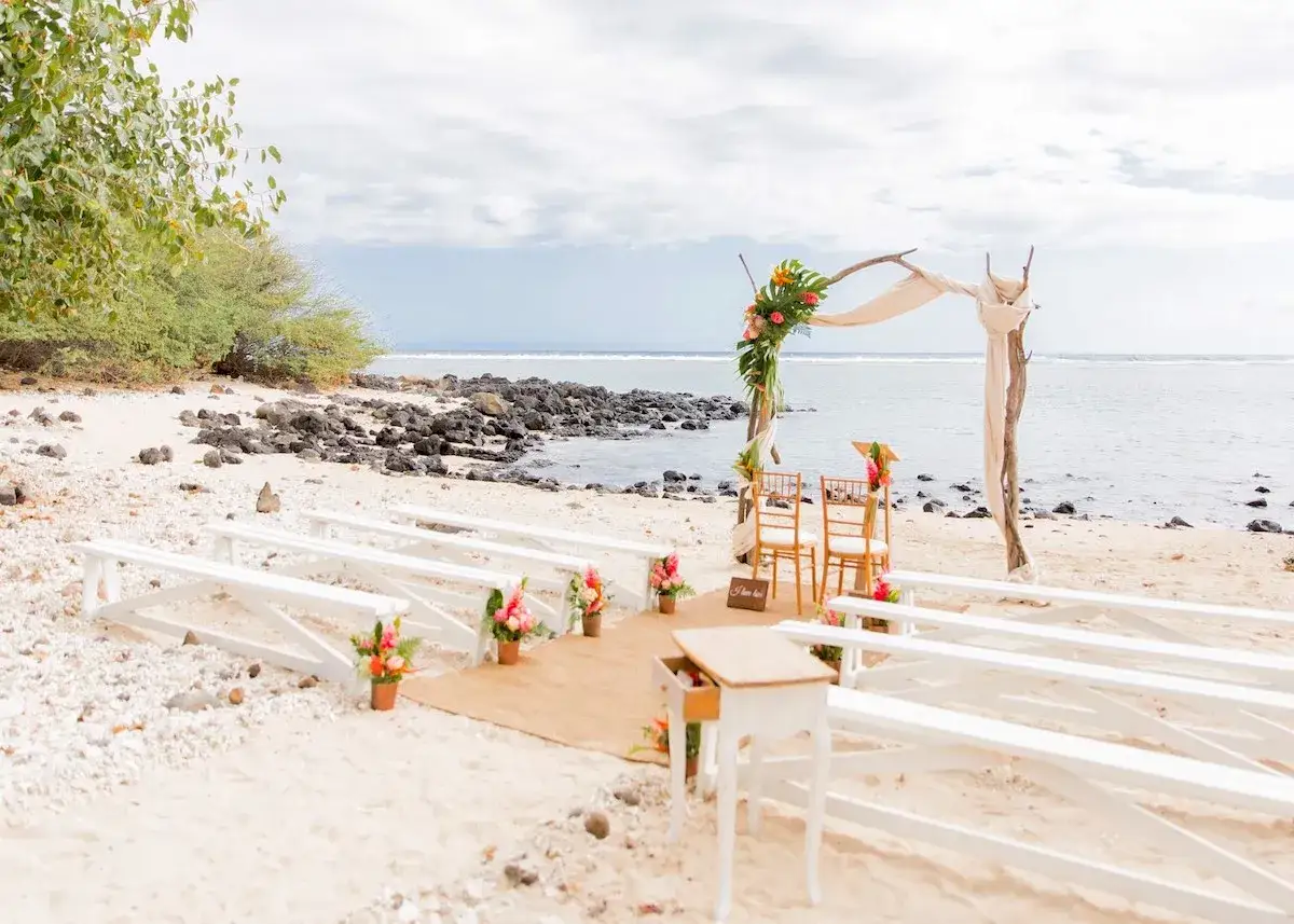 Allées de bancs blancs sur une plage de sable, décorées de fleurs tropicales, pour un mariage thème marin. L'arche de cérémonie est ornée de verdure et de fleurs colorées, face à l'océan.