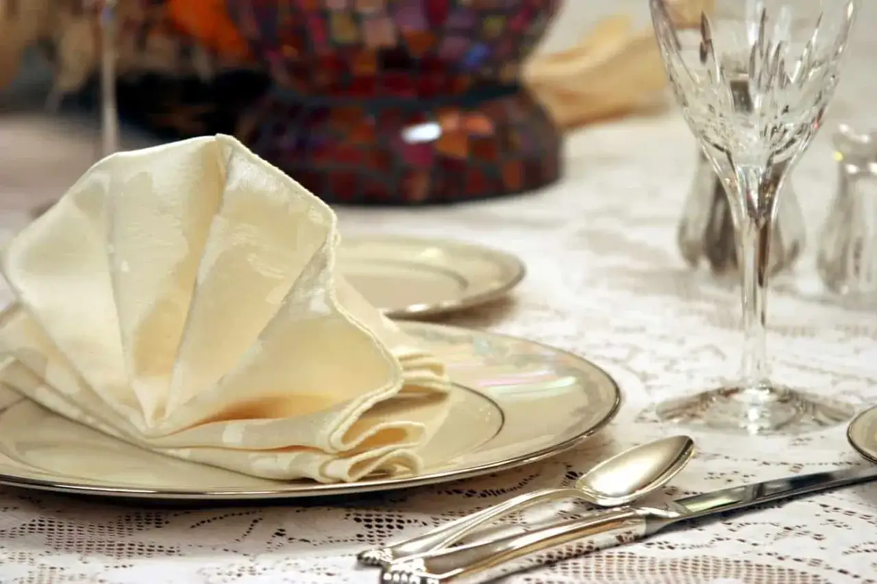 Un élégant modèle serviette de table en tissu crème, plié en éventail sur une assiette nacrée. La table est dressée avec des couverts en argent et un verre à pied.