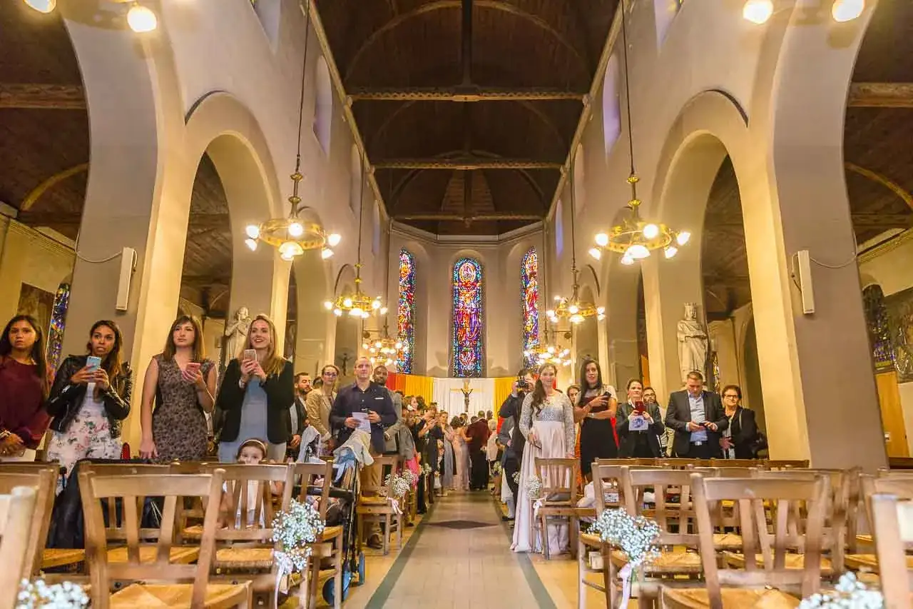 Des invit&eacute;s attendent dans une &eacute;glise, certains tenant leur t&eacute;l&eacute;phone, pour le cortege eglise mariage. Les vitraux illuminent la nef.