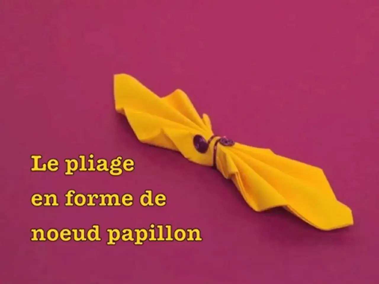 Une serviette jaune pli&eacute;e en forme de noeud papillon, tenue par un anneau. Un pliage &eacute;l&eacute;gant pour une table festive.