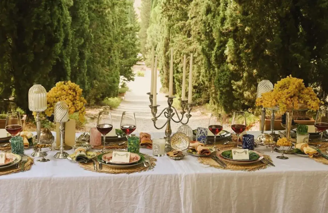 Une table dressée en extérieur, sous des cyprès, pour un protocole fiançailles. Vins rouges, bougies, fleurs jaunes et vaisselle élégante invitent à la célébration.