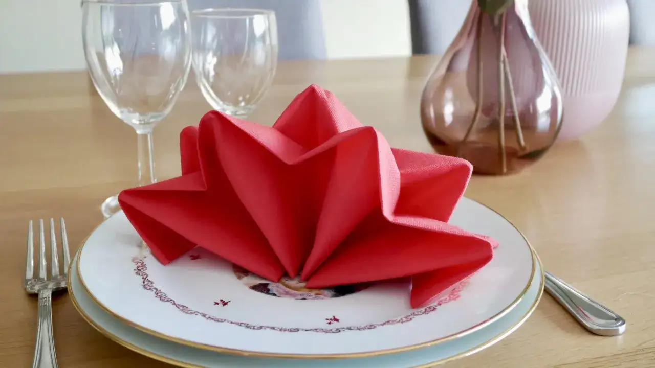 Une belle serviette rouge pliée en étoile pour un pliage serviette nouvel an festif. Assiette décorée, verres et couverts sur une table en bois.