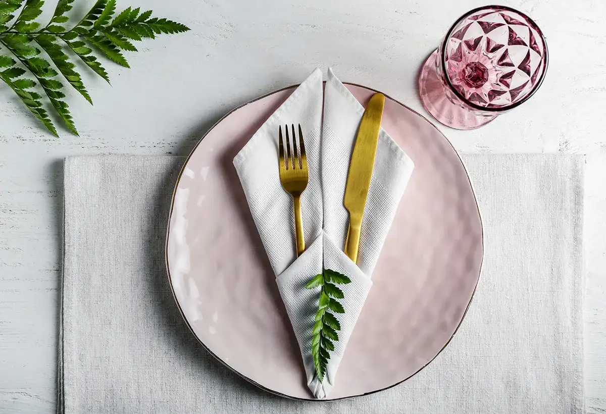 Un élégant pliage de serviette papier dans une assiette rose, ornée d'une feuille de fougère et de couverts dorés. Une touche de nature pour une table raffinée.