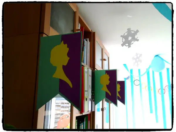 Guirlande "décoration reine des neiges à faire soi même" avec silhouettes jaunes sur fond violet et vert, suspendue devant une bibliothèque.
