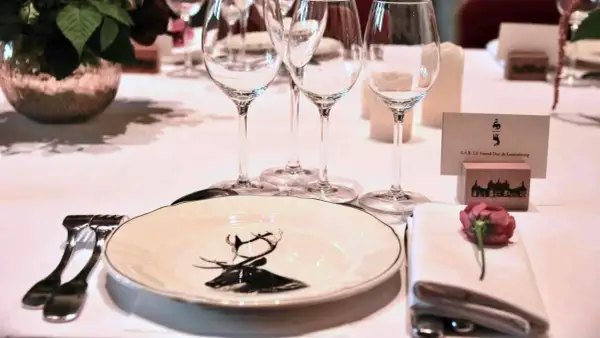 Une table dressée pour un dîner de gala, avec une assiette ornée d'un cerf, des verres à vin et une carte de visite "S.A.R. LE Grand-Duc de Luxembourg". La fourchette à la française est prête pour le festin.