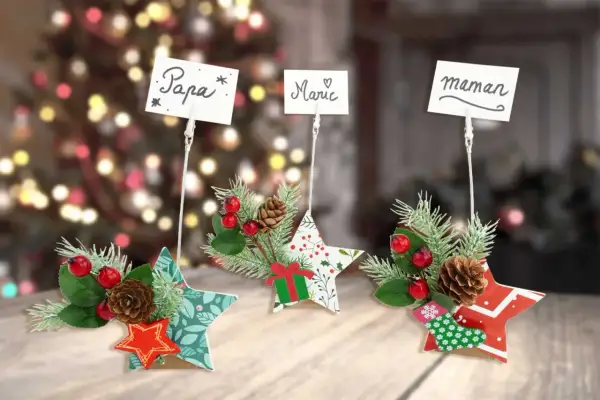 Porte nom original à faire soi-même : étoiles décorées de branches de sapin, baies rouges et pommes de pin, avec des noms écrits sur des cartes.
