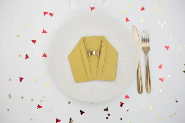 Pliage serviette nouvel an en forme de veste, orné d'un nœud papillon doré, sur une assiette blanche. Confettis sapins et étoiles rouges et dorés.