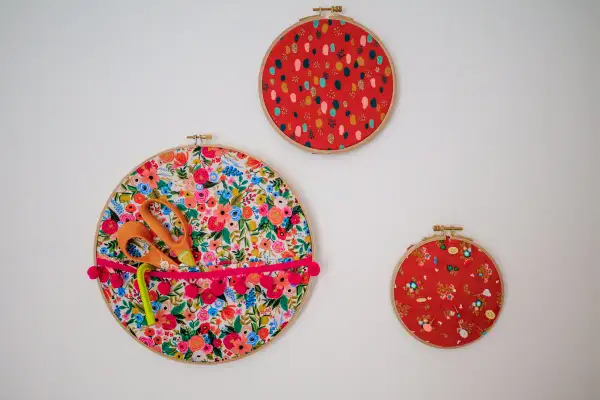 Décoration murale avec trois cercles à broder. Un grand cercle fleuri sert de rangement pour des ciseaux et un stylo, orné d'un ruban à pompons. Deux autres cercles rouges décoratifs complètent l'ensemble.