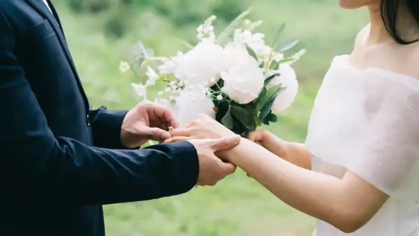 Un homme en costume met une bague à la main d'une femme, bouquet de fleurs blanches. Un poème mariage Baudelaire pour un amour éternel.