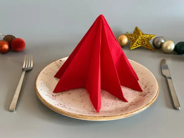 Un sapin en serviette papier rouge décore une assiette, prêt pour un repas festif. Des boules de Noël et une étoile dorée complètent la mise en scène.