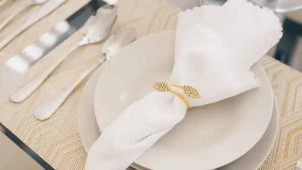 Une serviette blanche mariage, ornée d'un anneau doré scintillant, repose sur une assiette. Des couverts argentés sont disposés à côté.