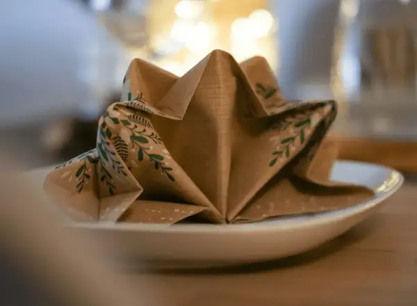 Une serviette en papier marron avec des motifs végétaux verts est pliée en forme d'étoile sur une assiette blanche. Ce pliage serviette triangle ajoute une touche festive.