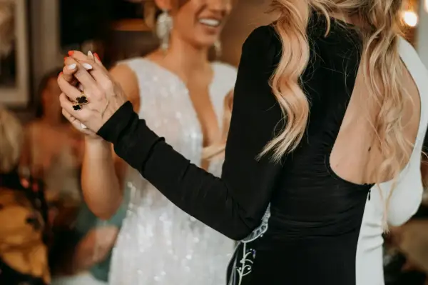 Deux femmes dansent, l'une en robe blanche scintillante, l'autre en robe noire. Elles se tiennent la main, un geste pour faire honneur au mariage.