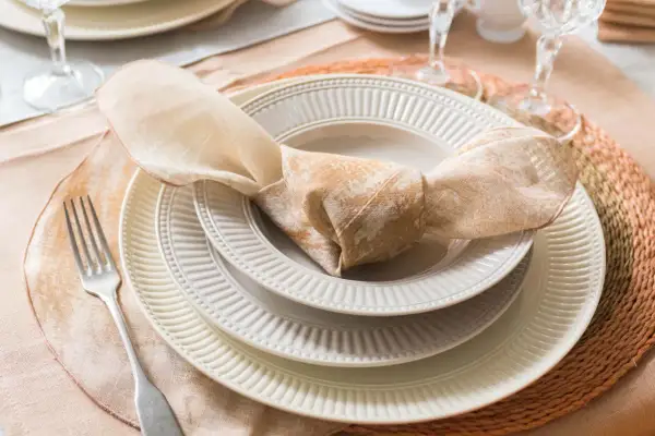 Une table dressée avec des assiettes empilées, une serviette en tissu nouée, une fourchette et des verres. La différence entre faïence et porcelaine est subtile, mais la qualité est évidente.