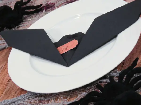 Une serviette noire pliée en forme de chauve-souris sur une assiette blanche, avec une étiquette "Joy". Décoration d'Halloween.
