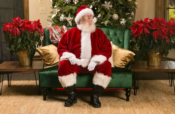 Père Noël, avec son déguisement de Noël fait maison, est assis sur un canapé vert, entouré de poinsettias rouges et d'un sapin décoré.